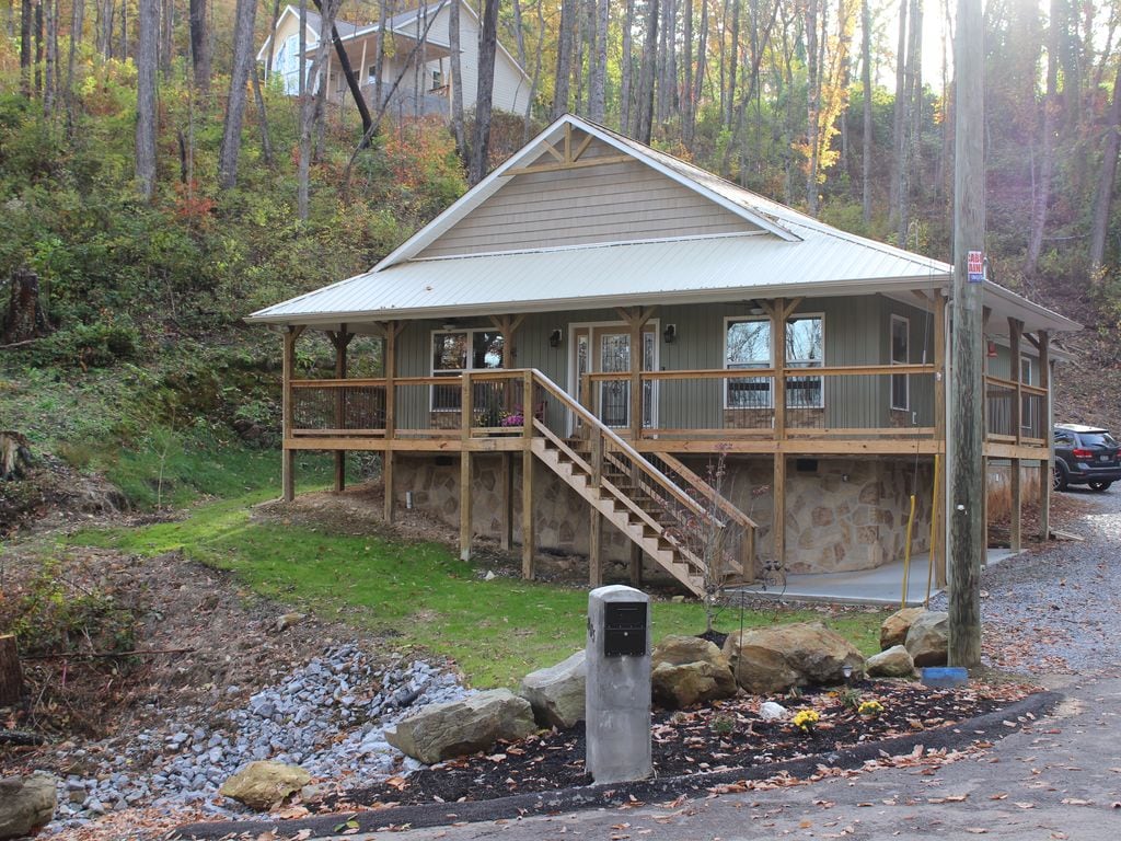A charming chalet exterior is presented, featuring a covered porch with wooden railings and stairs leading to the entrance. The building is surrounded by greenery, with a gravel path visible. Natural stone accents are included in the lower wall, and a car is parked nearby.