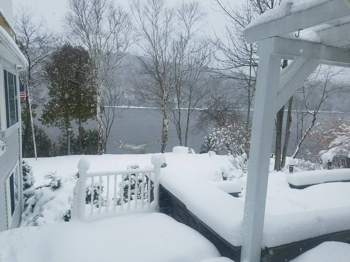 A serene winter scene showcases a snowy landscape surrounding the house. A layer of fresh snow blankets the patio area, with a view of the icy water in the background, framed by leafless trees and subtle hills. The atmosphere is enveloped in a quiet, wintry ambiance.
