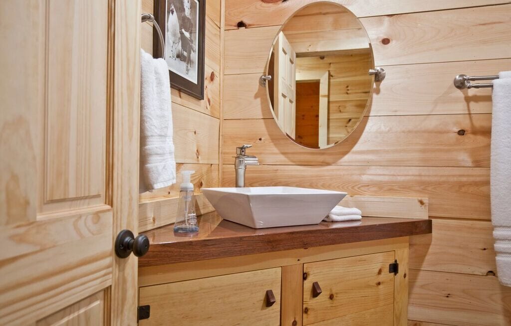 A wooden bathroom features light-toned shiplap walls and a round mirror above a modern, rectangular sink. Wooden cabinetry offers storage below, while neatly hung towels and a soap dispenser enhance functionality.
