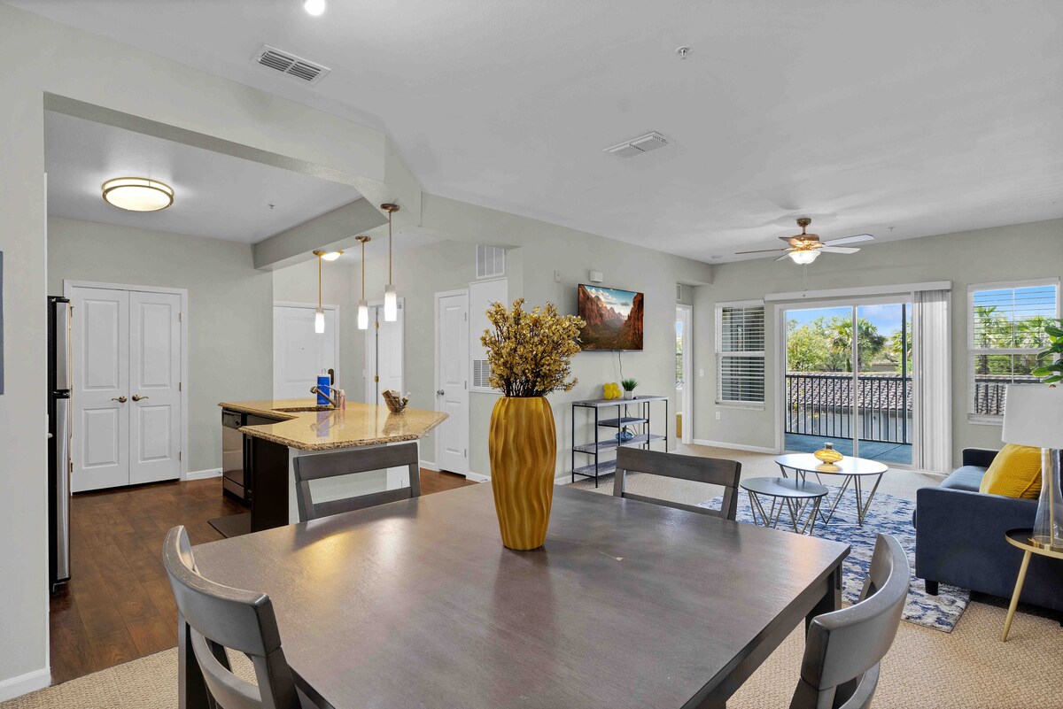 An inviting dining area is featured with a wooden table surrounded by four chairs. A bright vase with dried flowers adds a touch of decor. The open layout reveals a modern kitchen in the background and a comfortable living area with a sofa and coffee table, complemented by large windows offering natural light.