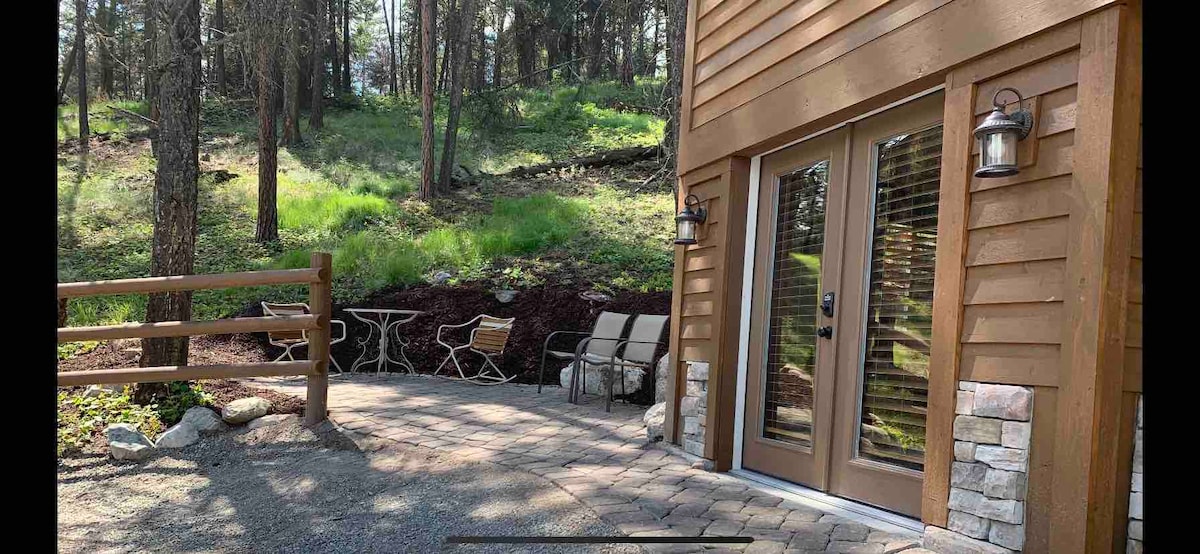 A private patio area is visible, featuring two lounge chairs and a small table nestled among natural greenery. The entrance to the cottage is framed by wood siding, with large glass doors providing a view of the outdoors. Nearby, the wooded landscape enhances the tranquil setting.