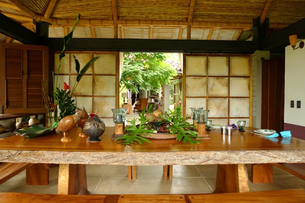 A large wooden dining table is set with various decorative items and plants. Natural light fills the space, highlighting the bamboo ceiling and the surrounding greenery visible through an open doorway. The warm tones of the wood blend harmoniously with the earthy decor.
