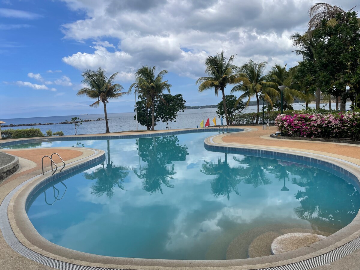 A tranquil pool area is showcased, with clear blue water reflecting the sky. Lush palm trees and vibrant floral arrangements frame the space, while the ocean is visible in the background. Colorful sailing flags add a cheerful touch to the serene setting.
