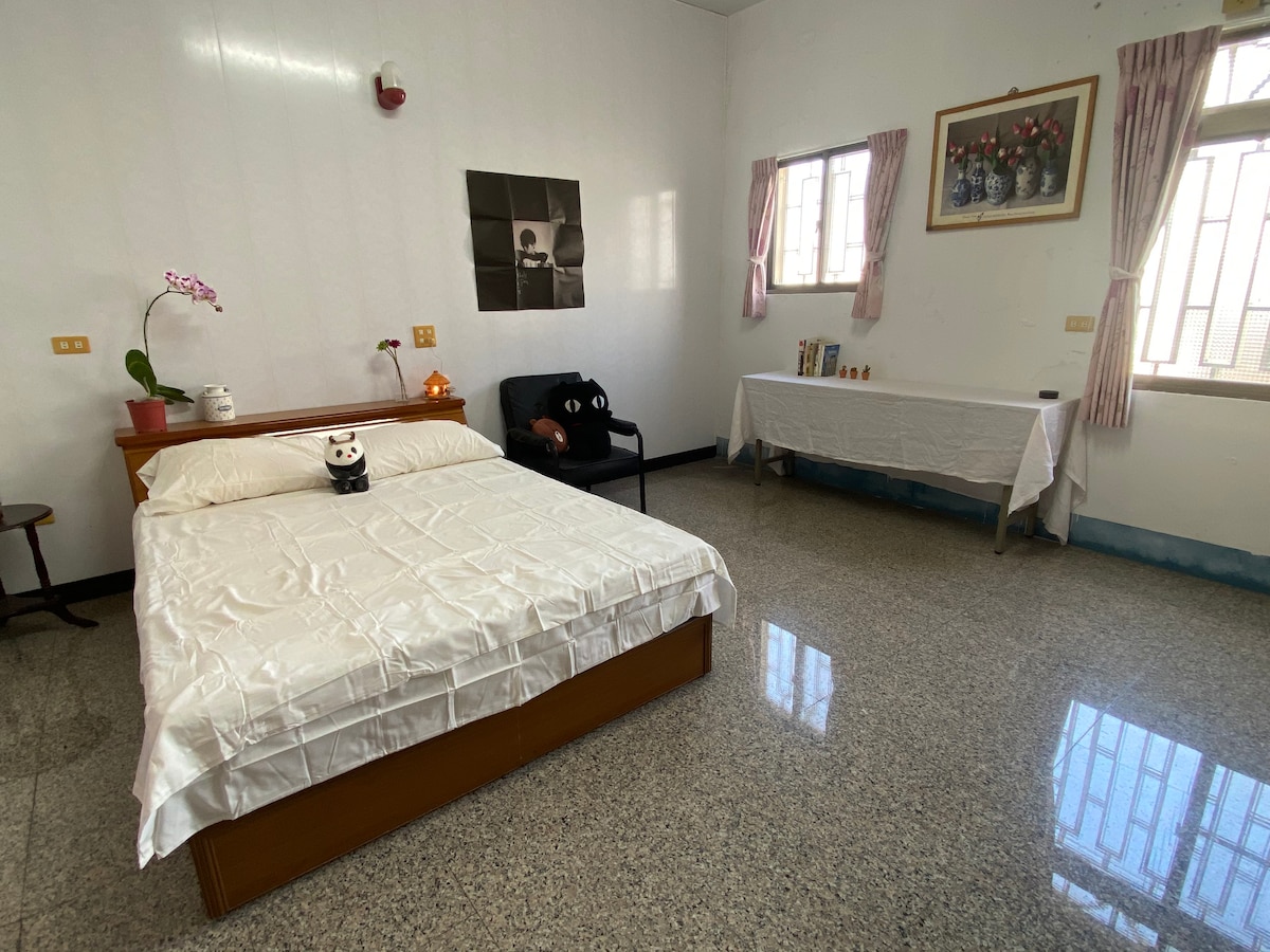 A bright room features a spacious bed with a white coverlet and decorative pillows. A small table with a vase of flowers sits beside the bed. A black armchair is positioned in one corner. Natural light filters through two windows adorned with light curtains.