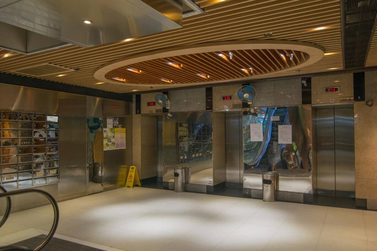 An entrance area is displayed featuring multiple elevators marked with lights above each door. The floor is tiled in a light color, and a caution sign is positioned on the floor. Clean, metallic walls reflect light, contributing to a bright environment.