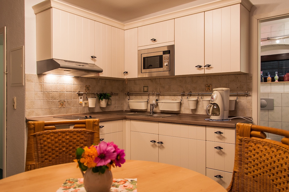 A modern kitchen is presented with white cabinetry and wood countertops. Cooking appliances, including a microwave and coffee maker, are visible. A round table with a fresh floral centerpiece is surrounded by wicker chairs, creating a cozy dining area.