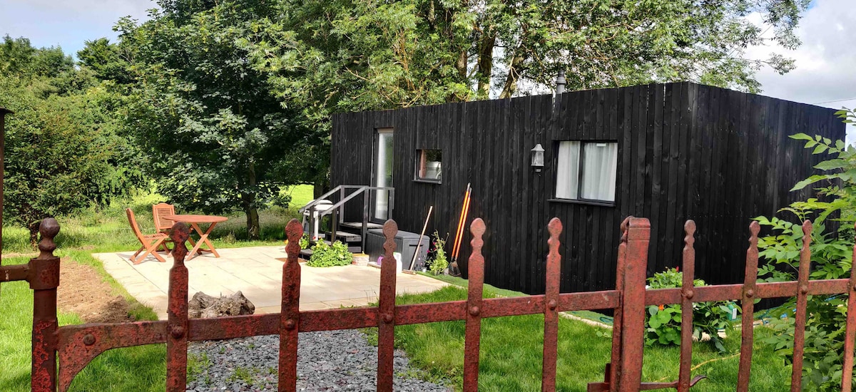 A modern black cabin is framed by lush greenery, featuring a small patio with outdoor furniture. A rustic iron gate adds character to the entrance, complemented by gravel pathways and a well-maintained lawn that enhances the natural surroundings.