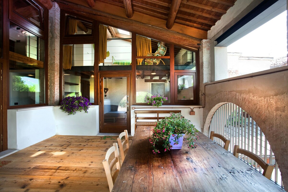 A spacious covered terrace features a wooden table surrounded by chairs, creating an inviting dining area. Lush potted plants add touches of greenery. Large windows allow natural light to fill the space while offering views of the tranquil surroundings, complemented by rustic wooden beams overhead.