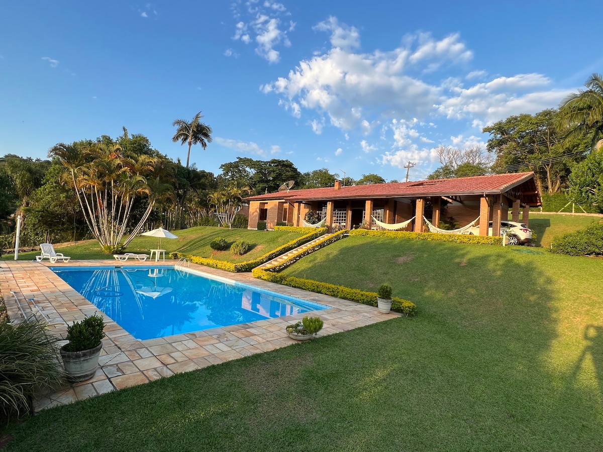 A spacious single-story house is set against a backdrop of lush greenery, featuring a welcoming patio area and a clear blue swimming pool. The exterior is enhanced by landscaped gardens and a terrace with hammocks, ideal for relaxing outdoors.