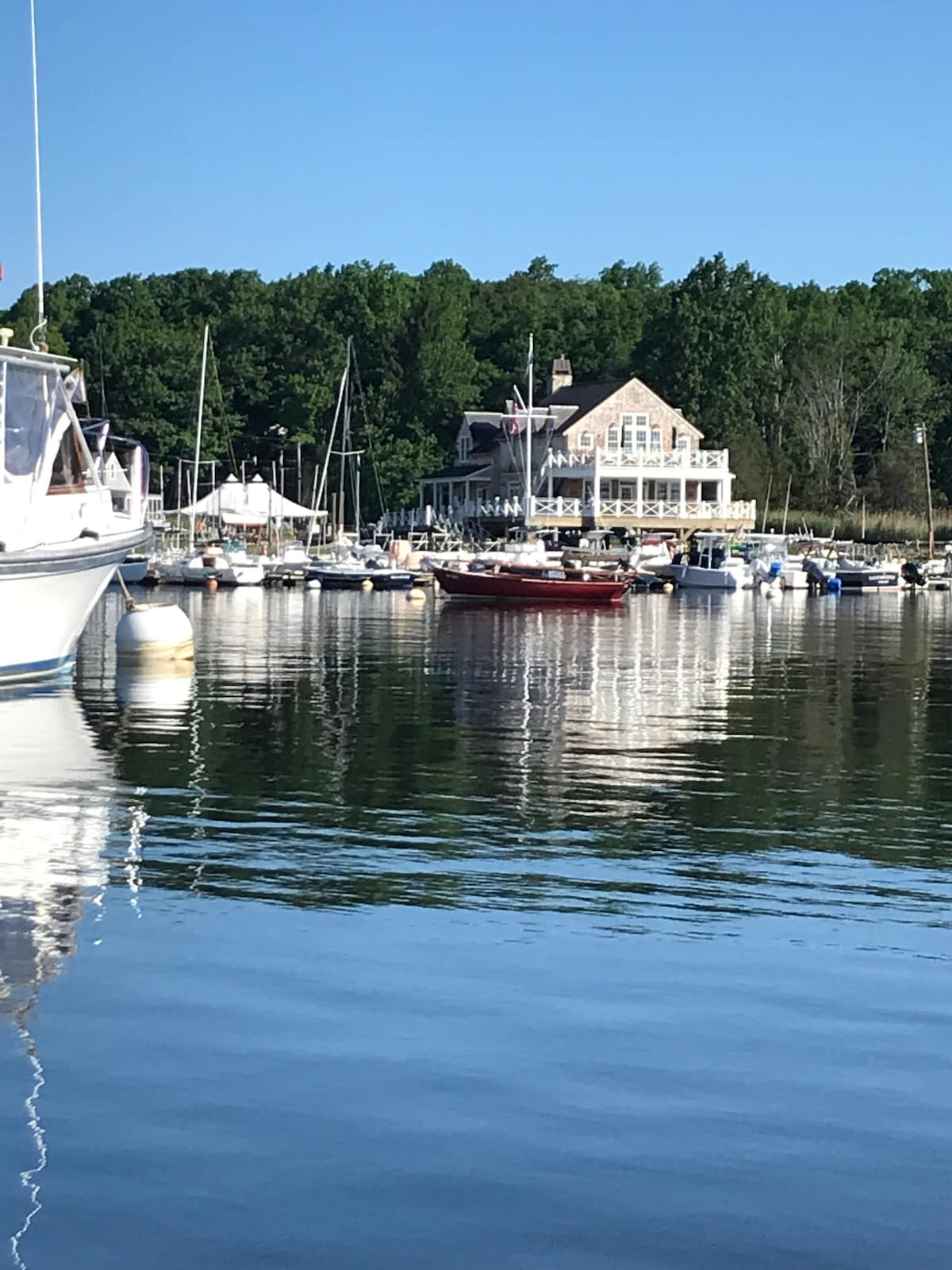 A peaceful harbor scene is captured, featuring various boats gently bobbing on calm waters. A charming multi-level house, framed by trees, overlooks the marina, reflecting in the water. The blue sky adds to the tranquil ambiance, enhancing the coastal setting.
