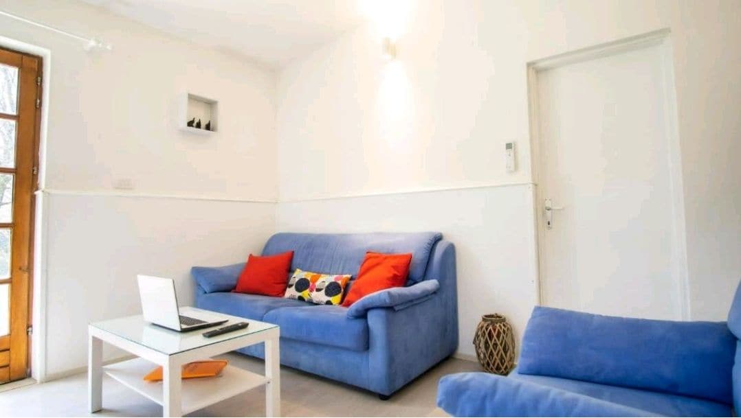 A bright living area features a light blue sofa adorned with colorful pillows and a matching armchair. A glass coffee table sits in front of the sofa, accompanied by a laptop. A wooden door leads outside, and natural light fills the space.