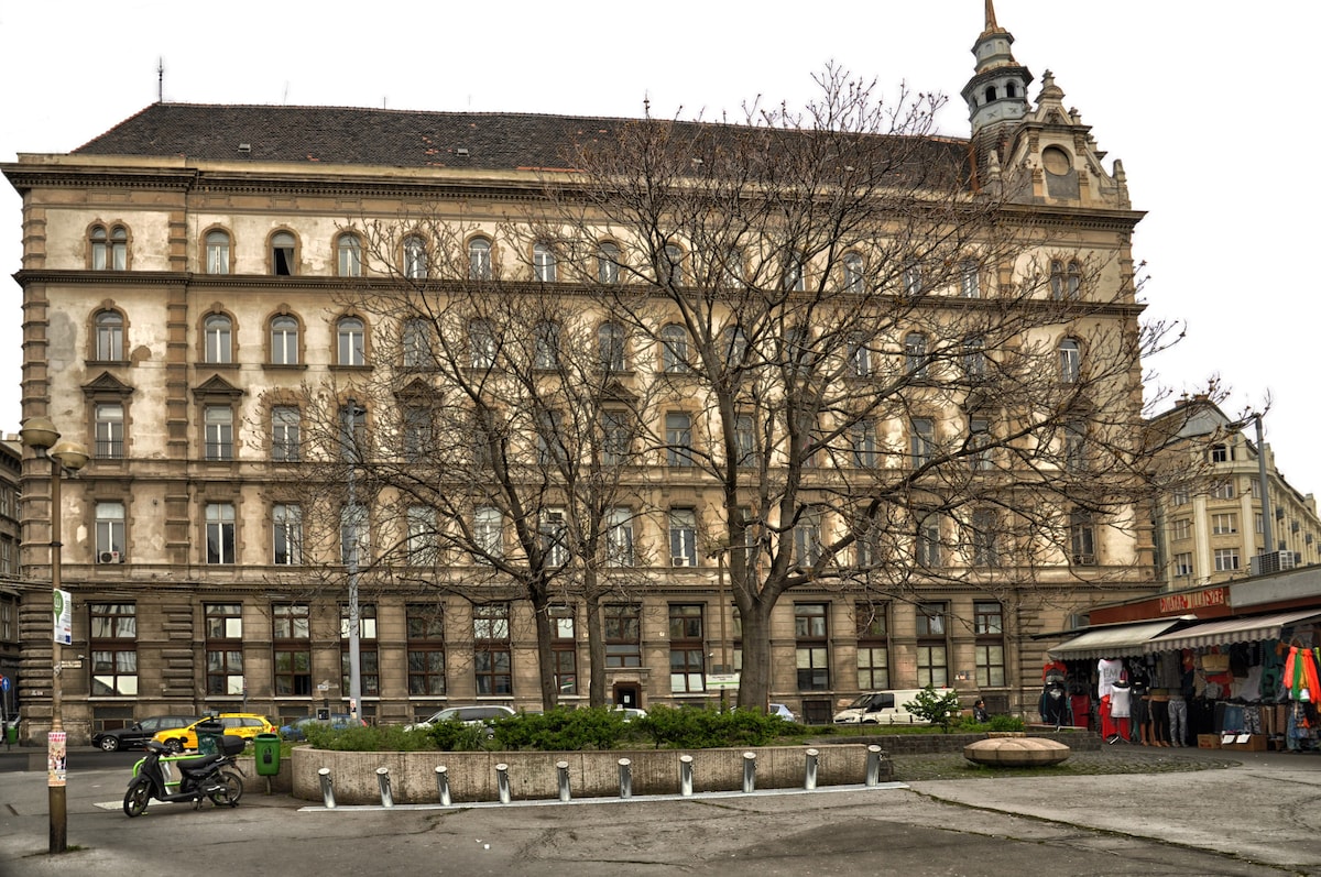A historic building stands prominently in the city center, featuring ornate architectural details. Large windows illuminate the facade, while a leafless tree adds natural elements to the foreground. Various sidewalk activities are visible, contributing to the vibrant urban atmosphere.