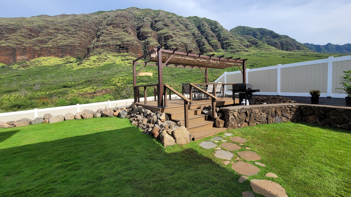 A spacious backyard deck is surrounded by lush green grass and features a wooden pergola. Stone pathways lead through the yard, with mountain views visible in the background. The area is enclosed by a white fence, offering both privacy and a natural setting.
