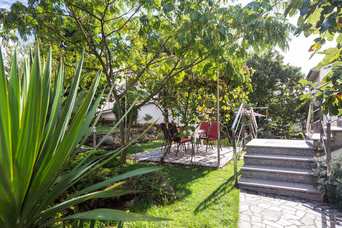 A private garden area features a set of red seats beneath leafy trees, providing shade and relaxation. Stone steps lead to the entrance of the apartment. Lush greenery surrounds the space, adding to the tranquil environment.