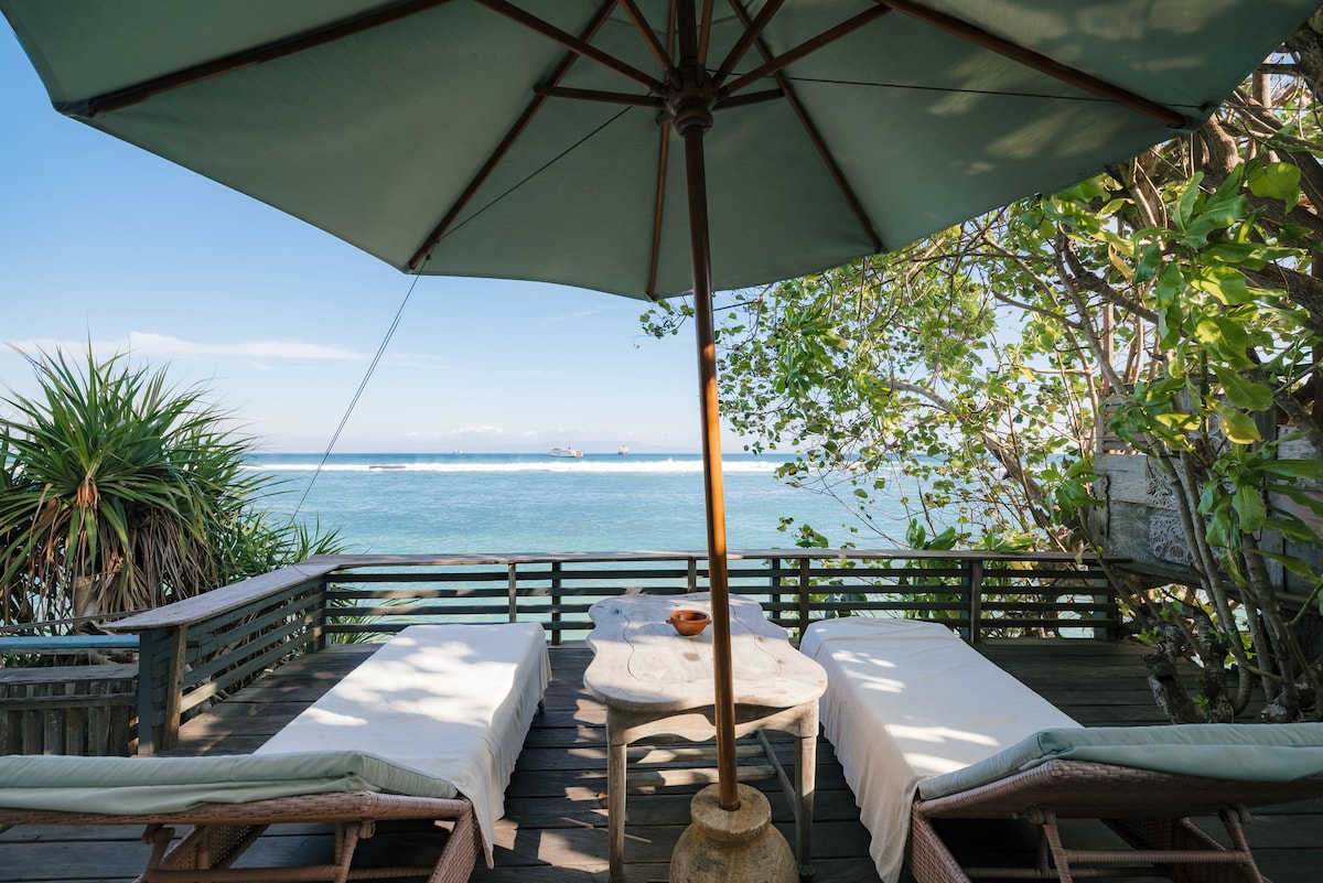 A sunny beachfront deck is equipped with two lounge chairs and a circular wooden table. A large umbrella provides shade, and tropical plants frame the area, while the ocean sparkles in the background, offering a serene view.