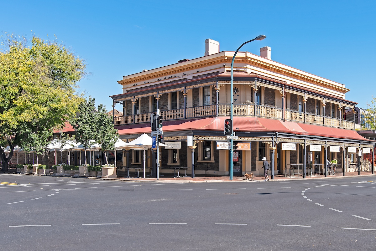 An elegant, two-story building features a traditional facade with a prominent balcony on the upper level. The corner location provides a view of a tree-lined street, while outdoor seating is visible, inviting a relaxed atmosphere for patrons.