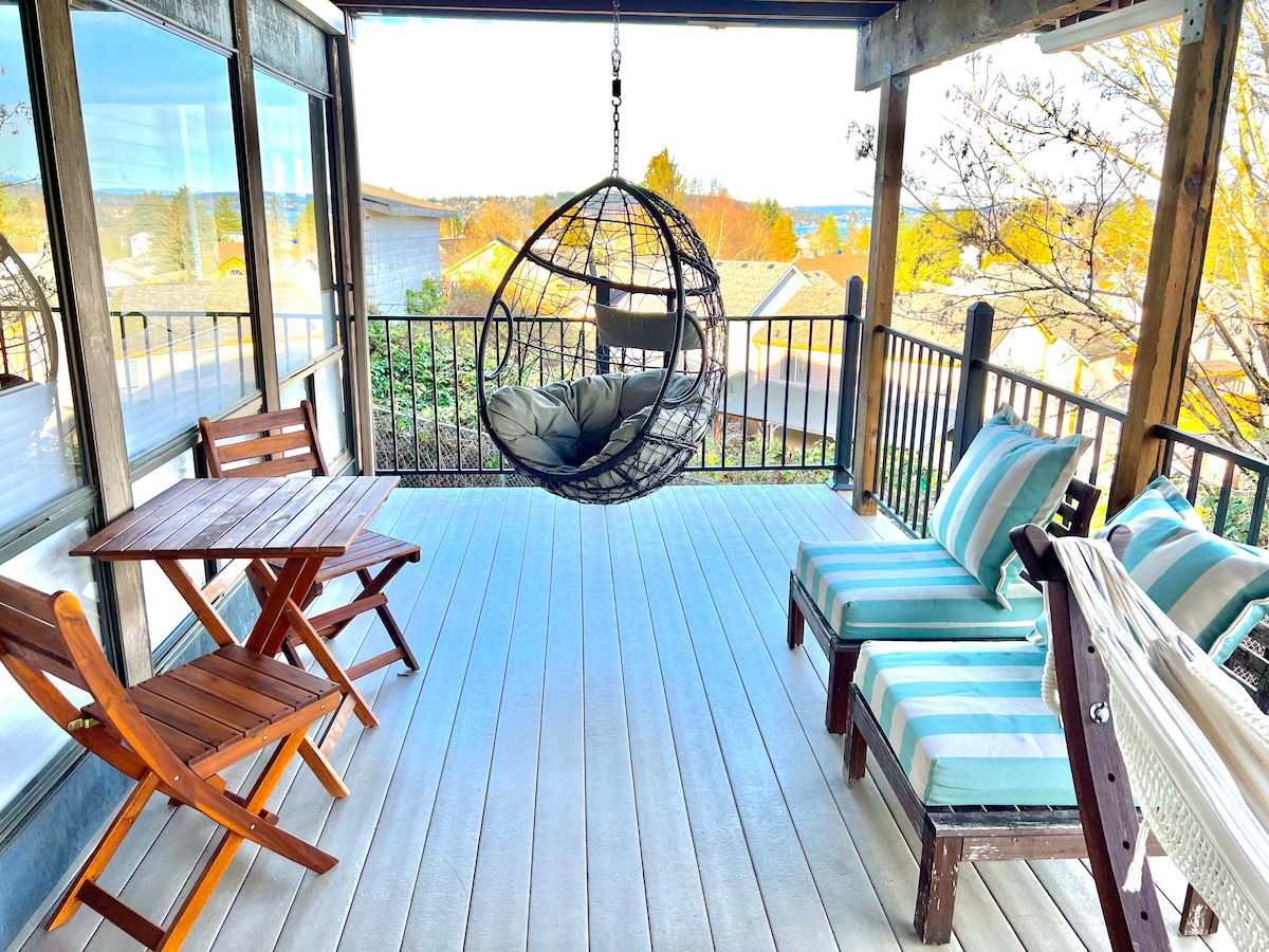 A shaded patio area features a hanging chair and a small wooden table with two folding chairs. Two lounge chairs with striped cushions are positioned nearby. Lush greenery and distant views of Lake Washington and the Cascade Mountains are visible through large windows.