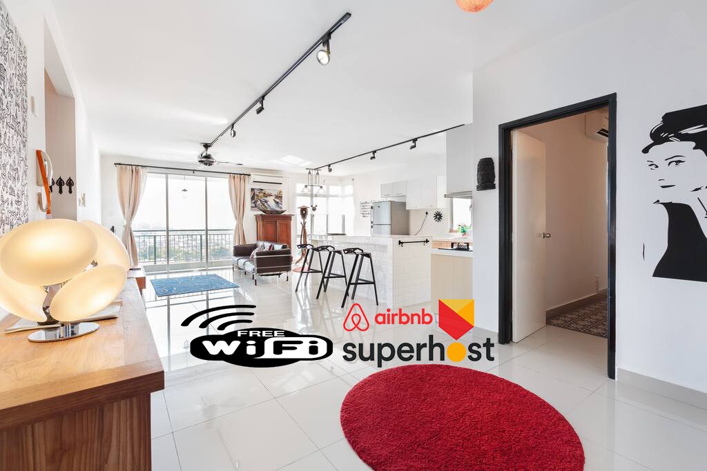 An open living space features a modern kitchen with sleek bar stools and a dining area. Natural light floods the room through large windows, revealing a scenic view. Decorative elements such as a vibrant area rug and wall art enhance the contemporary design.