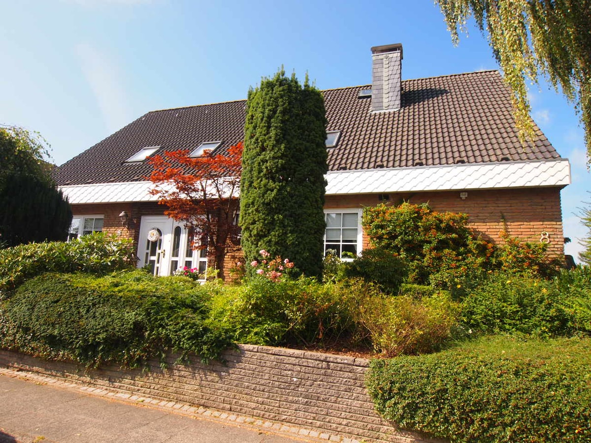 A modern brick house is showcased with a sloped roof and large windows. Lush greenery surrounds the property, including well-maintained bushes and a small tree. A pathway leads to the front entrance, enhancing the welcoming appearance of the home.