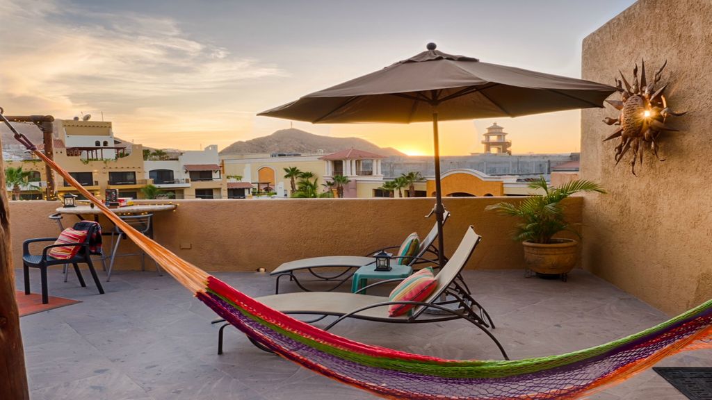A rooftop terrace is shown with a colorful hammock and a lounge chair positioned under a parasol. Views of a distant hillside and a sunset sky are visible. A table with a grill completes the inviting outdoor space.