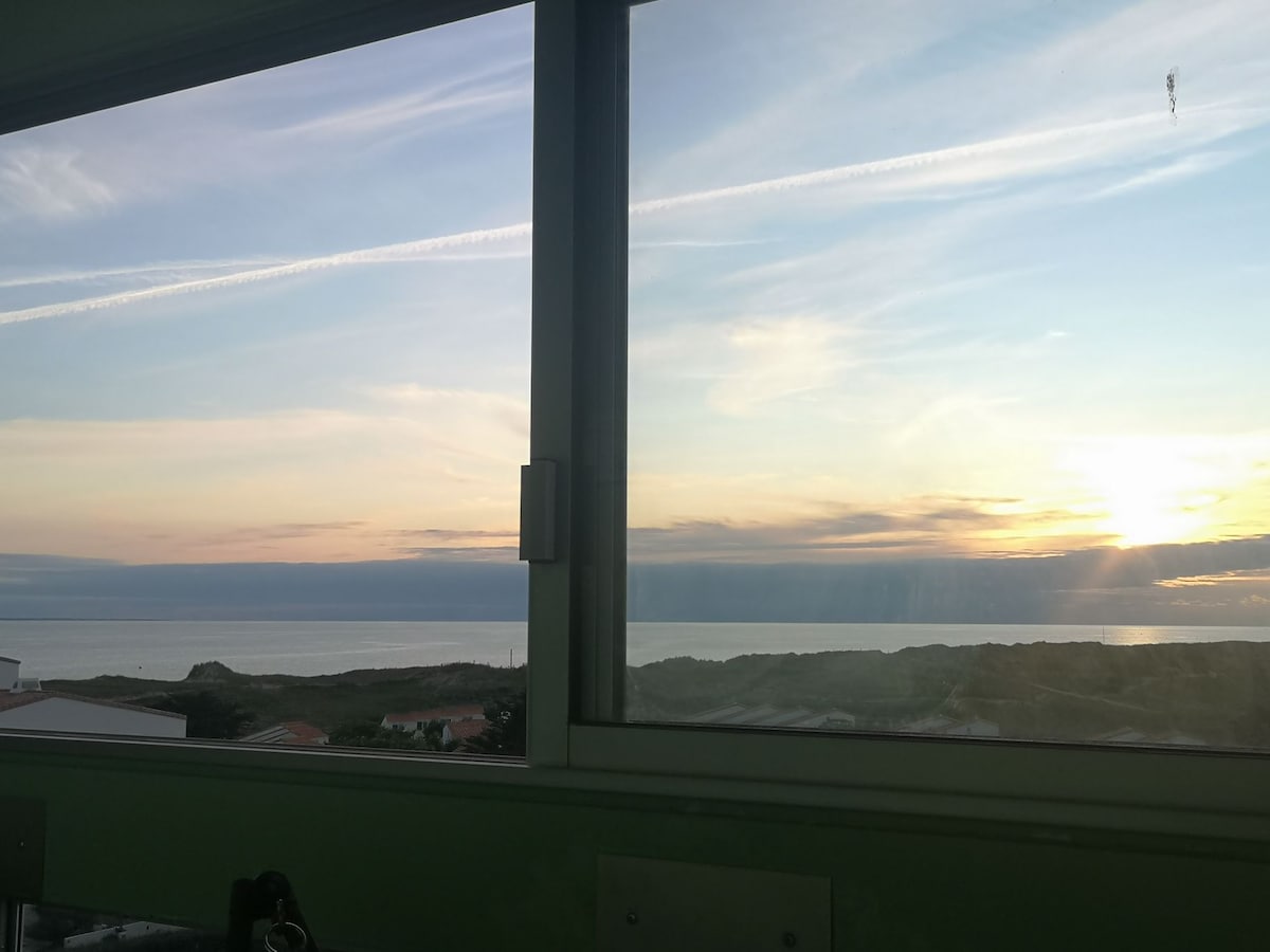 A large window frames a serene sunset over the sea, with soft hues of orange and pink reflecting on the calm water. The distant dunes are visible against the horizon, enhancing the peaceful coastal view.