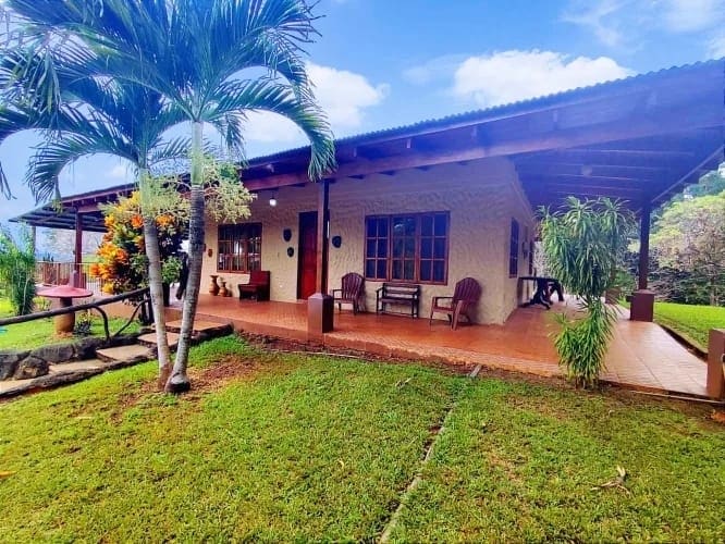 A spacious exterior view of the house reveals a covered terrace adorned with wooden seating and potted plants. Lush green grass surrounds the property, and tropical trees provide shade, creating a serene environment. The sky is bright, complementing the inviting scene.