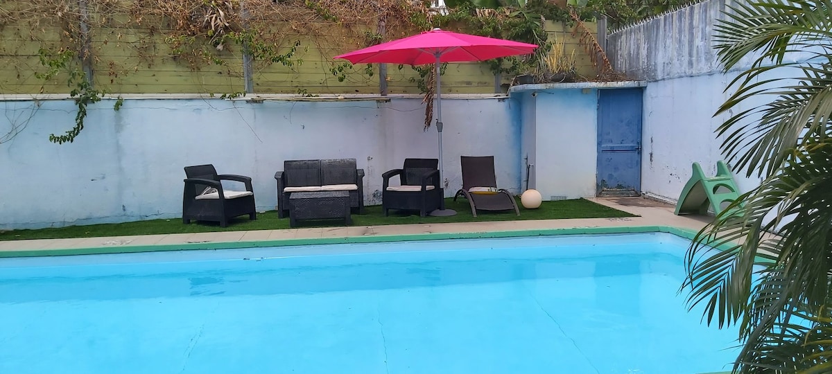 A refreshing pool is surrounded by a well-kept patio area, featuring several seating options, including lounge chairs and a shaded area with a bright pink umbrella. Lush greenery and a simple wall provide a sense of privacy and relaxation.