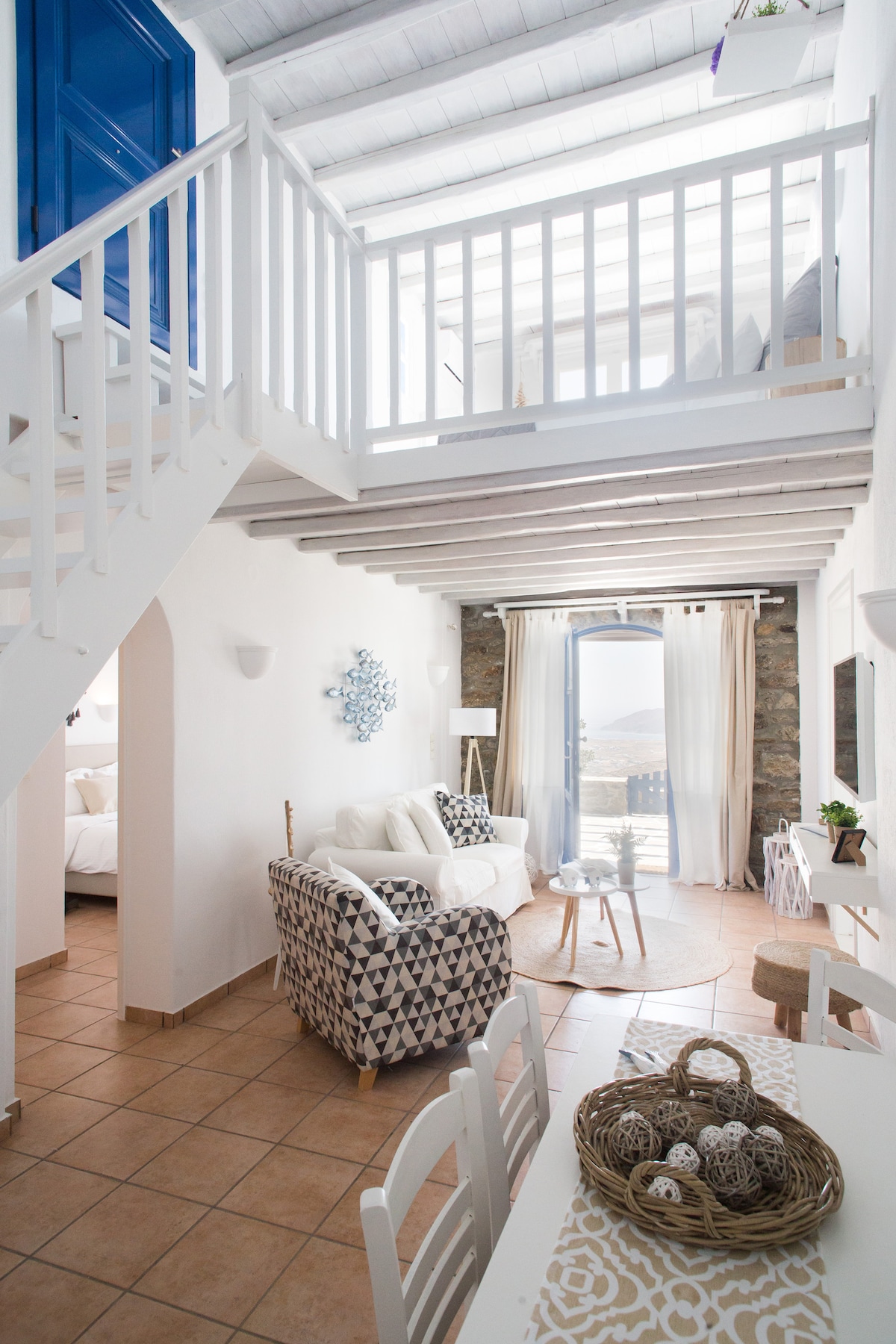 An open-plan living area features a cozy seating arrangement with a patterned sofa and a circular rug. A staircase leads to a lofted area, while large windows provide ample natural light. The stone wall enhances the unique ambiance, complemented by neutral decor elements.