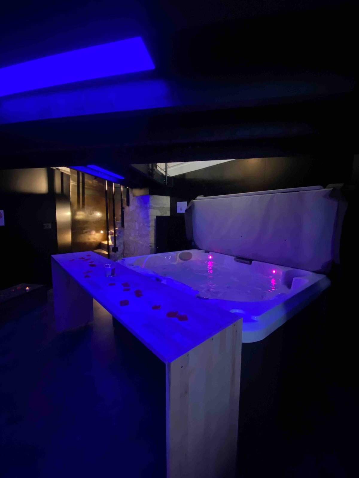 A private jacuzzi is highlighted in a dimly lit space, with purple and blue lighting creating a relaxed atmosphere. Decorative rose petals are scattered across the wooden edge, adding a touch of romance to the setting.