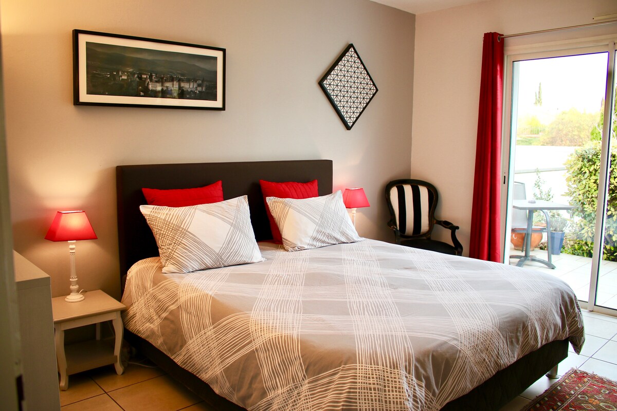 A cozy bedroom features a spacious bed with a patterned duvet and red accent pillows. A framed photograph adorns the wall, while a chair is positioned beside the bed. Soft red lamps provide ambient lighting, and glass doors lead to an outdoor terrace surrounded by greenery.