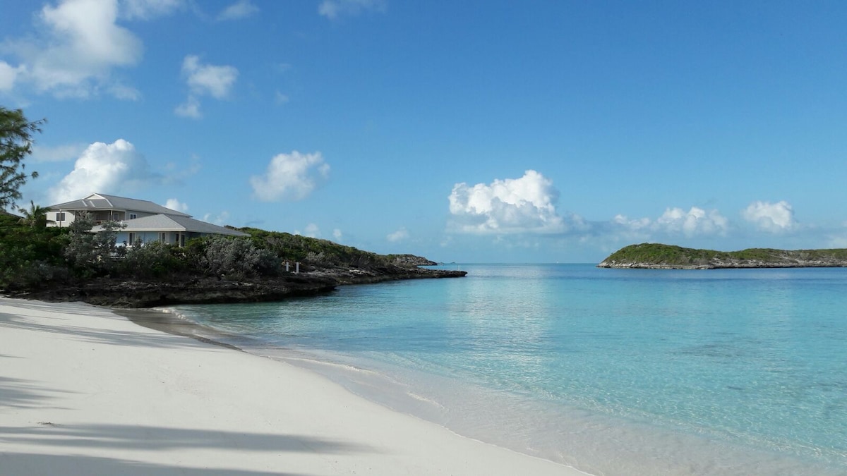 A serene view of a white sandy beach stretches towards calm turquoise waters. Soft waves gently lap at the shoreline, with the villa visible in the background against a clear blue sky dotted with fluffy clouds. Lush greenery frames the scene, adding to the tranquil ambiance.