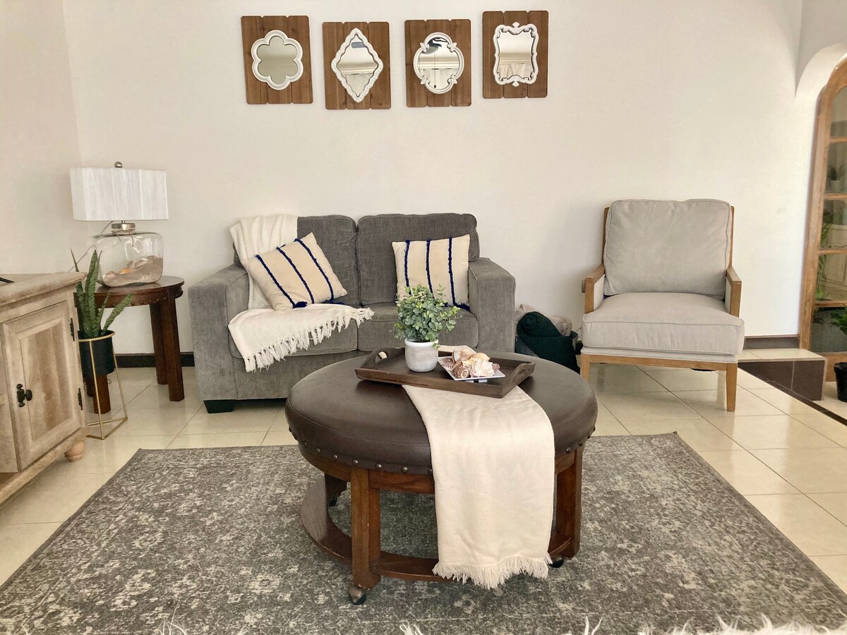 The living area features a gray sofa with decorative pillows and a cozy throw. A round coffee table holds a small plant and pastries, while a light-colored armchair provides additional seating. Framed mirrors are displayed above the sofa, adding visual interest to the space.