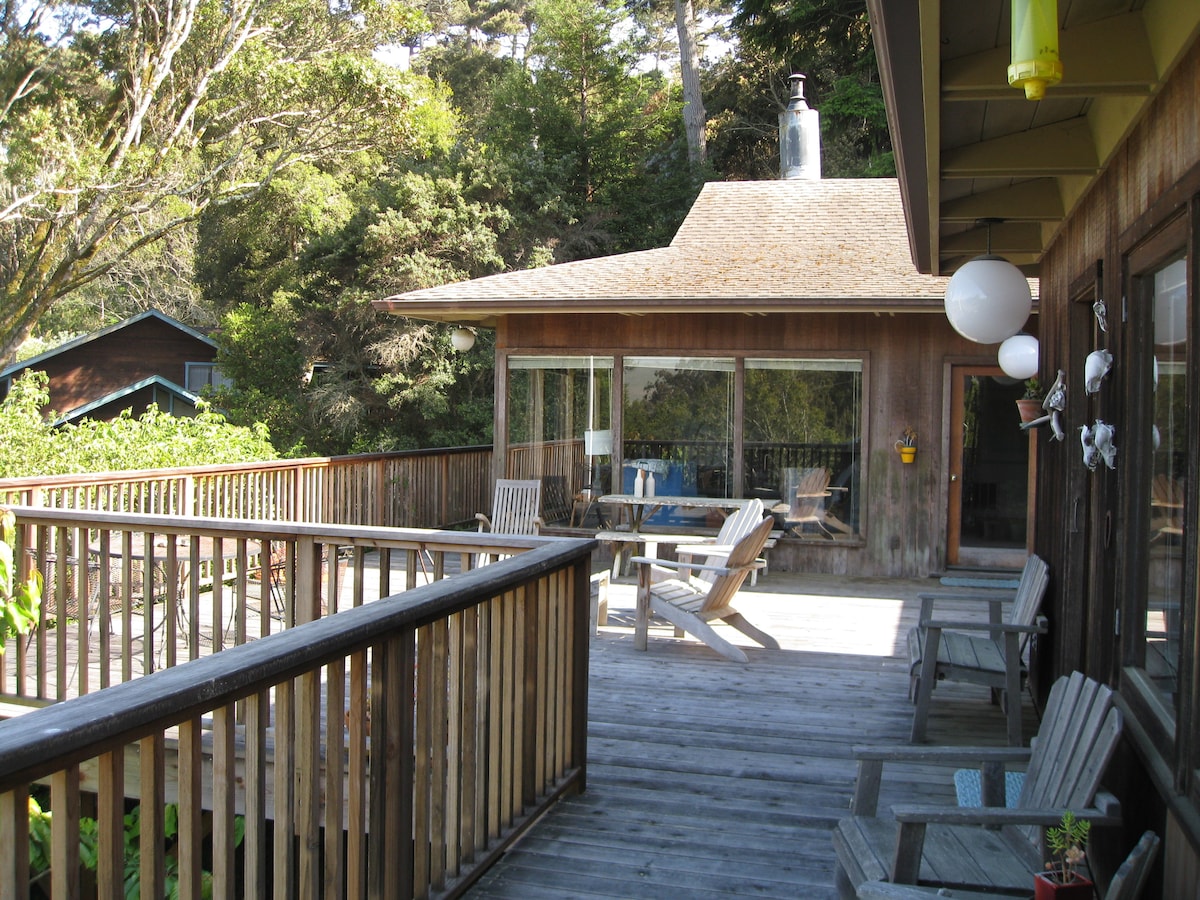 A spacious wooden deck is visible, lined with comfortable chairs and a table for outdoor dining. The cottage with large glass windows is situated in the background, surrounded by greenery, offering a serene setting for relaxation.