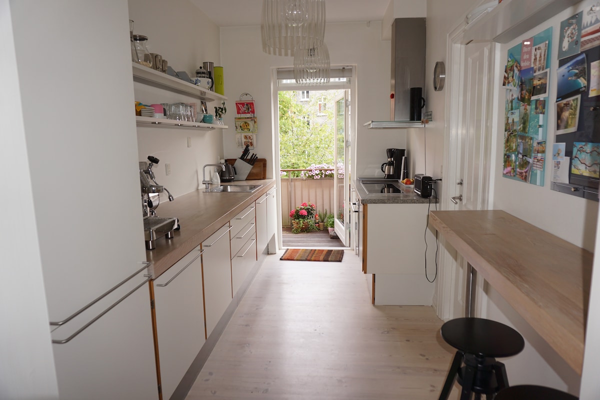 The kitchen features modern cabinetry and a spacious layout, with stainless steel appliances and a countertop under natural light. A doorway leads to a balcony adorned with blooming plants, enhancing the view of the outdoor area. A table and bar stool provide additional seating.