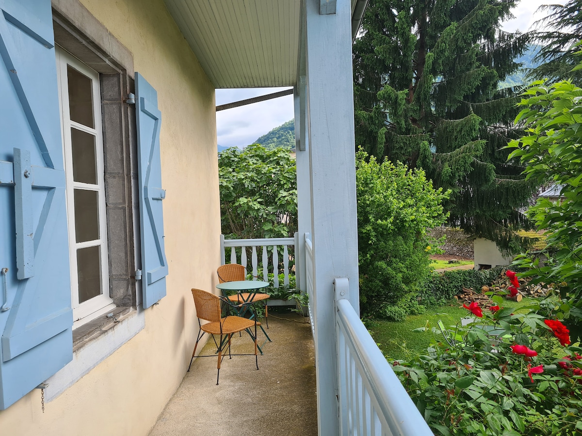 A balcony features a small round table accompanied by two wicker chairs, set against a backdrop of greenery. Blue shutters frame a window, and vibrant flowers are visible in the garden below, enhancing the setting's natural charm.