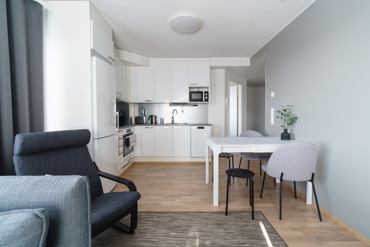 A modern kitchen area is visible, featuring white cabinetry and stainless steel appliances, including a refrigerator, oven, and microwave. A dining table with four chairs occupies the space, with a comfortable armchair positioned nearby. Warm wood flooring complements the neutral wall colors.