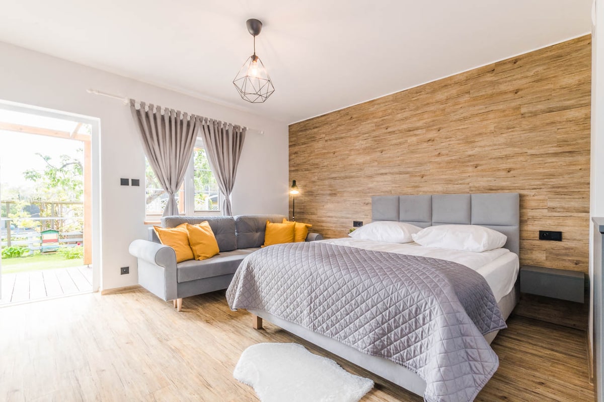 A spacious sleeping area features a large bed with white bedding and a quilted grey cover. A grey sofa with orange cushions is positioned nearby. Light enters through a window adorned with grey drapes, illuminating the wood-paneled wall and floor.