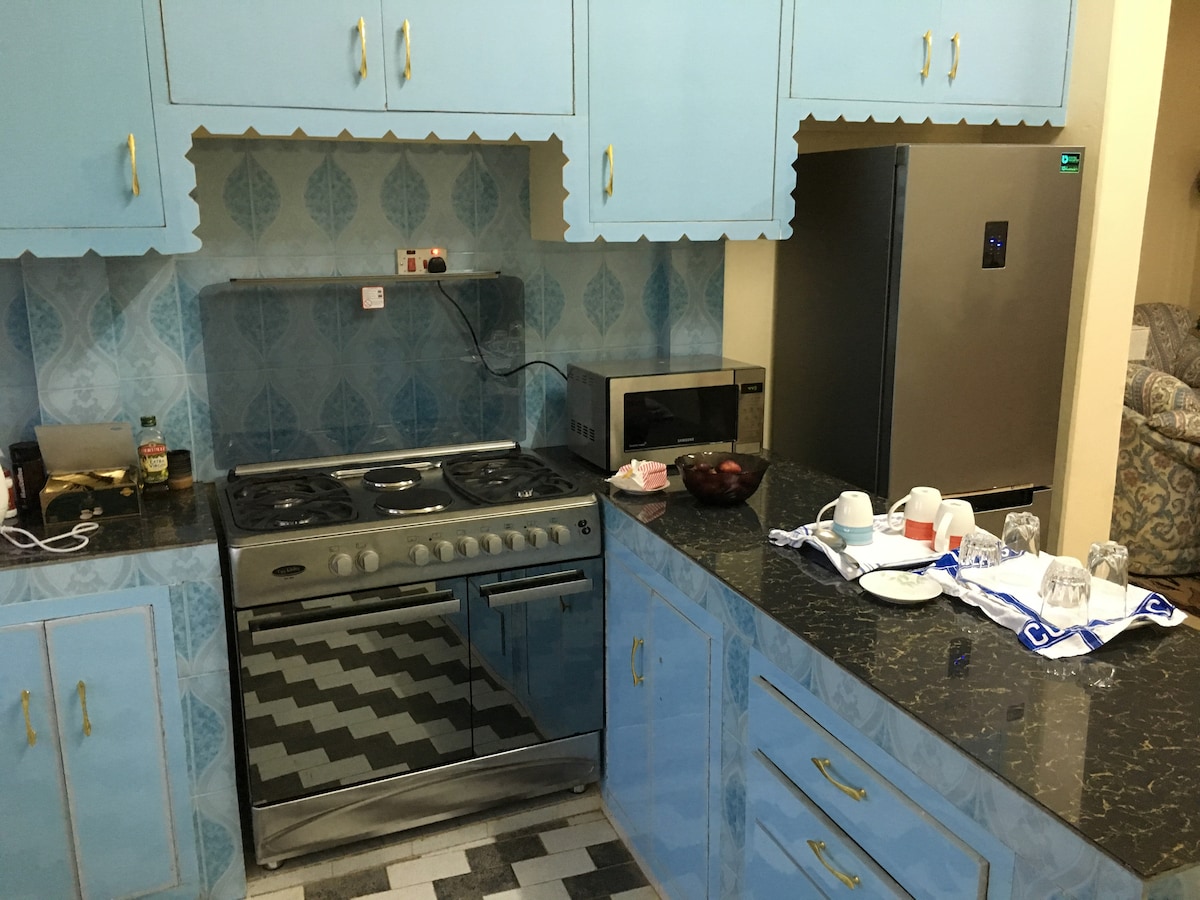 The kitchen features blue cabinetry, complemented by a marble countertop. A modern gas stove is positioned on one side, with a microwave and refrigerator for convenience. Various utensils and crockery are neatly arranged, providing a functional space for meal preparation.