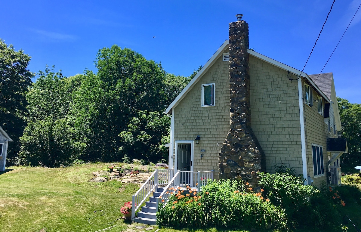 A charming cottage is presented with a stone chimney on the side. Lush greenery surrounds the home, complemented by colorful flowers in the landscaped garden. A set of steps leads to the entrance, with a clear blue sky above.