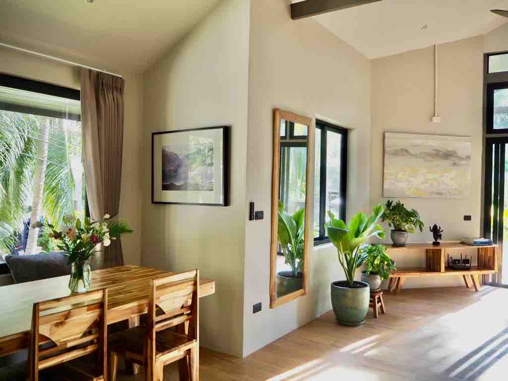 A bright living space is illuminated by natural light from large windows. A wooden dining table is surrounded by four chairs. Lush green plants are located throughout the area, complemented by contemporary artwork on the walls.