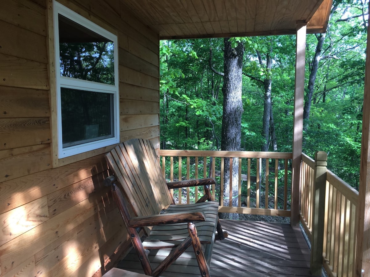 A cozy balcony features a wooden bench with wide armrests, positioned to overlook a lush wooded area. Natural light filters through the surrounding greenery, creating a serene outdoor seating space.