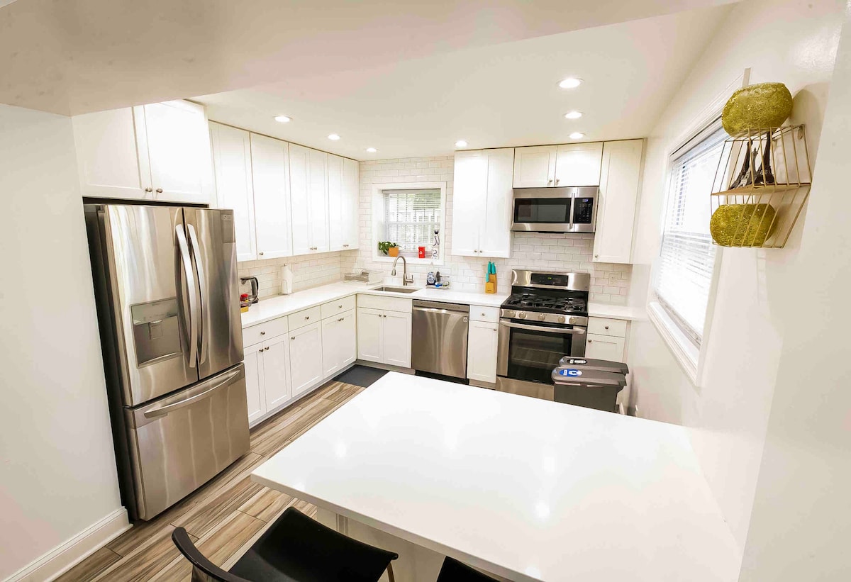 A modern kitchen is featured with stainless steel appliances, including a refrigerator and oven. White cabinetry complements the light-colored countertops. A spacious table is positioned at the center, surrounded by chairs. Natural light illuminates the area through a window, enhancing the clean and functional design.