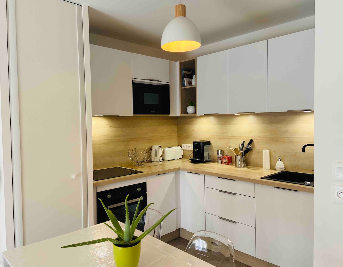 The kitchen is equipped with modern cabinetry and appliances, including an oven, microwave, and coffee maker. A light fixture hangs above a small dining table, while a potted plant adds a touch of greenery. Soft lighting enhances the inviting work surfaces.