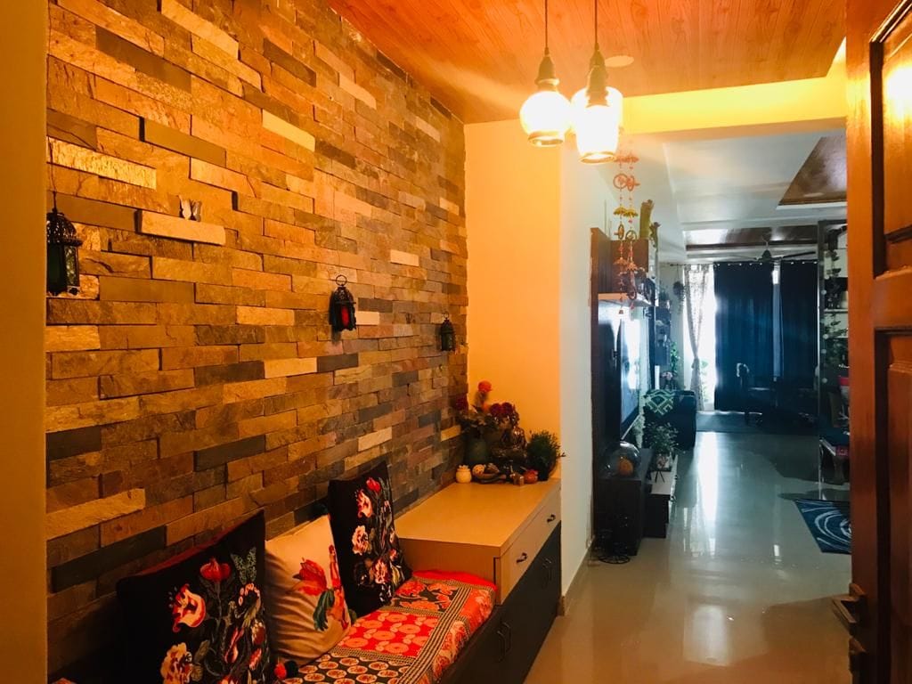 A cozy hallway features a stone accent wall, enhancing the natural aesthetic. There is a built-in seating area with colorful cushions, complemented by decorative lanterns. A continuation of the hallway leads to a living space with dark curtains and various plants, contributing to a warm ambiance.