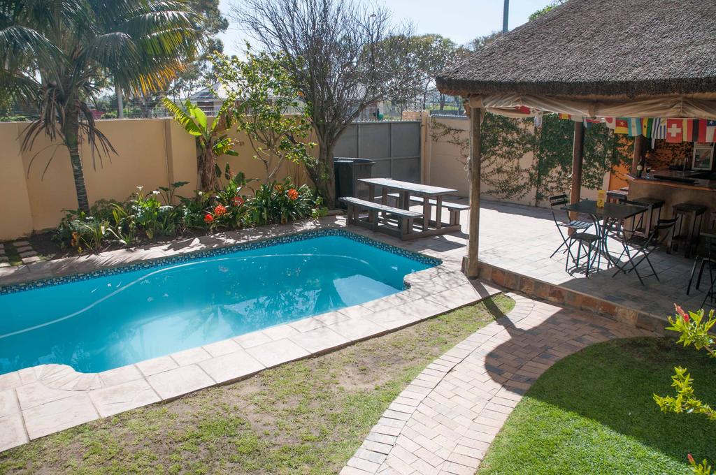 A private garden is featured, showcasing a clear blue pool surrounded by a stone patio and lush greenery. A thatched-roof gazebo with seating is visible, along with a picnic table nearby, offering an inviting space for relaxation and socializing.