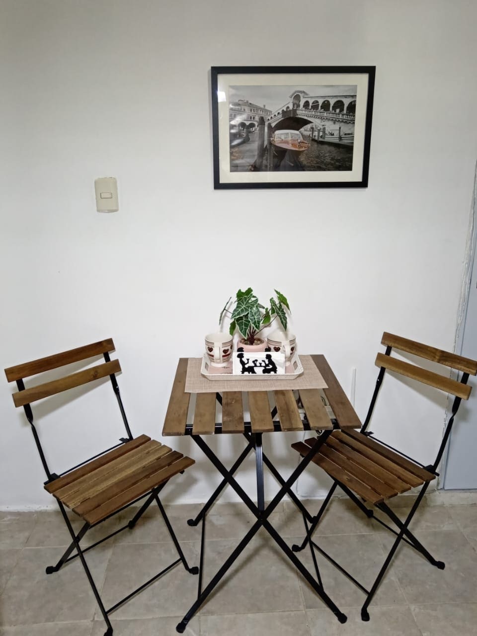 A small dining area is presented, featuring a wooden table surrounded by two folding chairs. A decorative plant sits at the center, with a neutral mat underneath. Above the table, a framed black-and-white photograph depicts a scene with boats, enhancing the minimalist design.