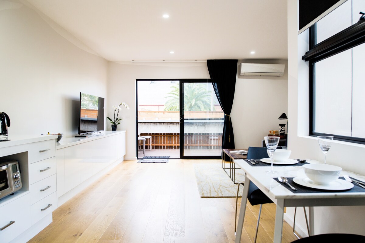 A bright living space is showcased, featuring a flat-screen TV and a small dining table set for two. Floor-to-ceiling windows allow natural light to fill the room, with glimpses of greenery visible outside. Minimalist decor complements the light wood flooring.