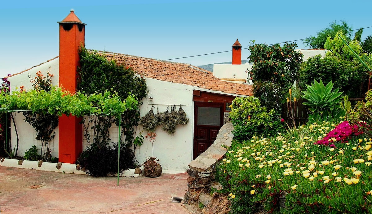 The exterior of a traditional Canarian finca is visible, featuring colorful flowering plants and lush greenery. The white-walled structure has a terracotta roof and a distinctive chimney. A variety of plants and vines surround the entrance, creating an inviting outdoor space.