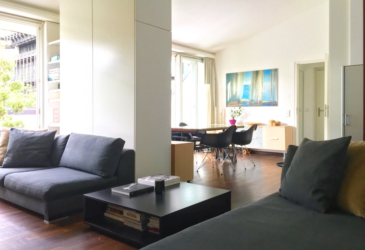 A modern living area features two dark sofas and a coffee table with neatly arranged magazines. A dining area with black chairs and a light wooden table is visible in the background. Bright natural light fills the space through large windows, complemented by a calming wall art piece.
