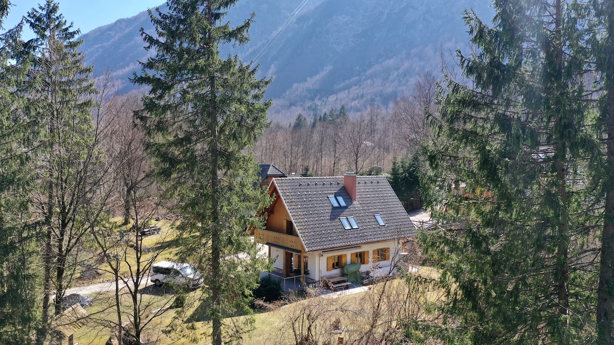 A two-story mountain home is surrounded by lush greenery, showcasing a classic wooden exterior with a sloped roof and multiple windows. The property features a covered porch and a spacious lawn, with mountain views visible in the background.